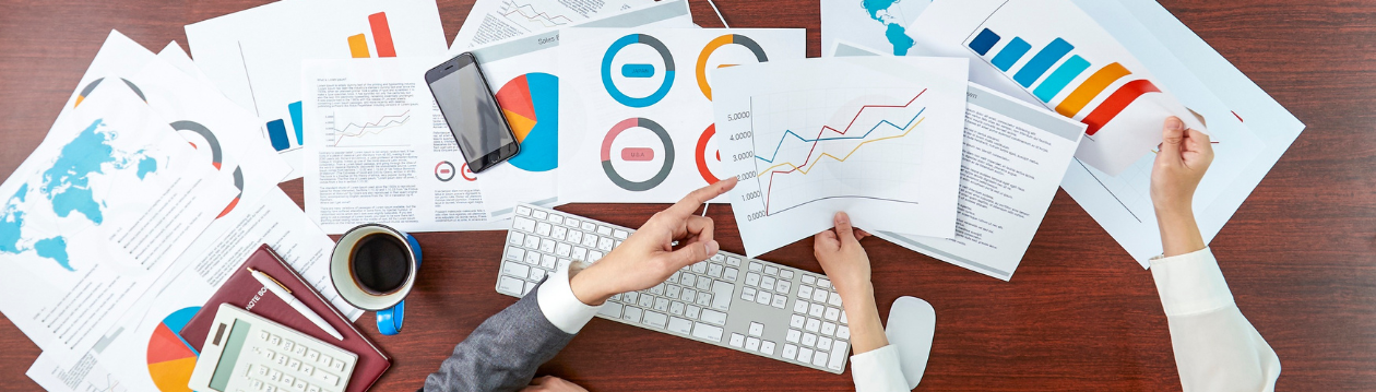 Schreibtisch mit Tastatur, Kaffee und Smartphone, umgeben von Diagrammen; zwei Personen zeigen auf Blätter mit Grafiken.