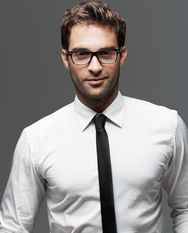 A brown-haired man with glasses wears a white shirt and a black tie, he smiles at the camera.