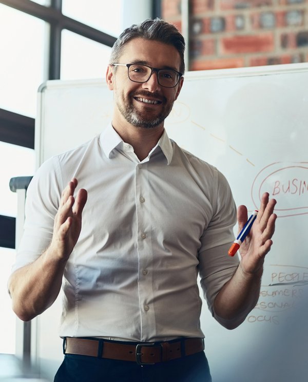 A grey-haired man with a white shirt and glasses stands in front of a whitboard. He is explaining something.