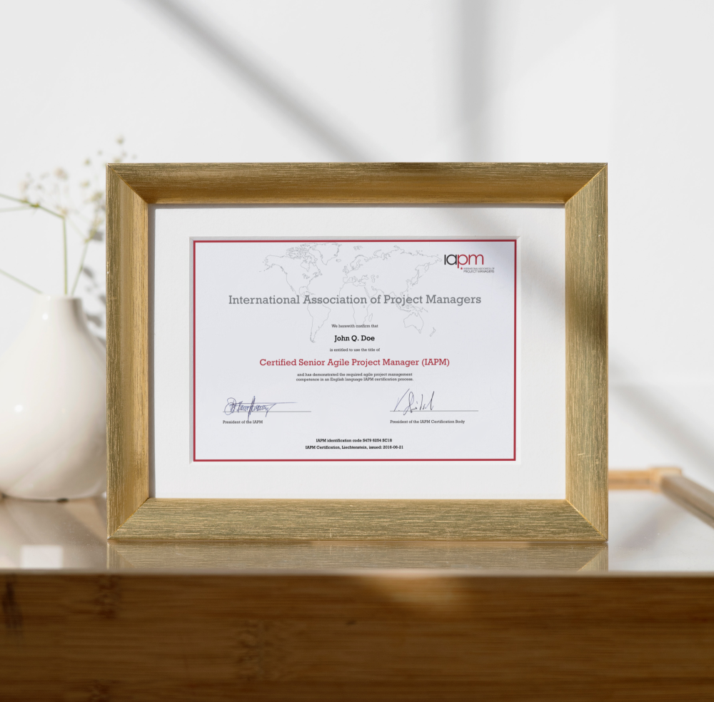 The Certified Senior Agile Project Manager certificate is in a wood-coloured picture frame and stands on a table. A flower can be seen in the background.