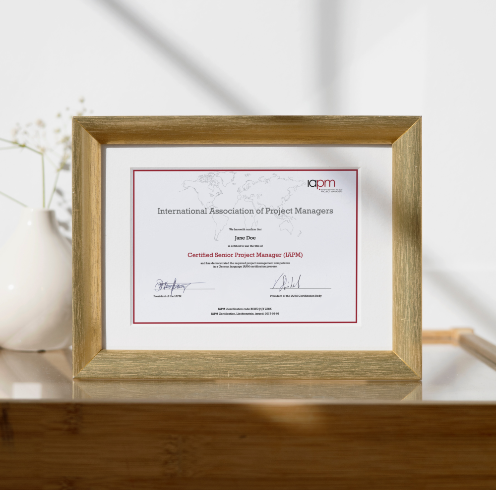 The Certified Senior Project Manager certificate is in a wood-coloured picture frame and stands on a table. A flower can be seen in the background.
