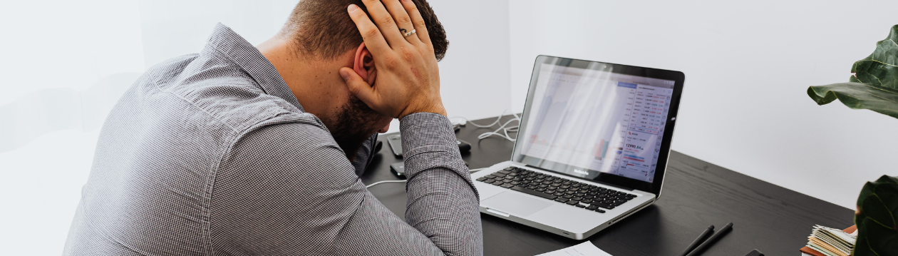 Ein Mann sitzt mit gesenktem Kopf und hält sich den Kopf vor einem Laptop mit Diagrammen am Schreibtisch.
