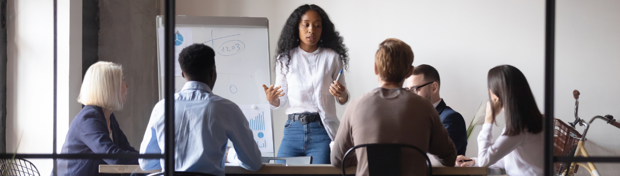 A woman presents to five people at a conference table with a flipchart and charts. A woman presents to five people at a conference table with a flipchart and charts.
