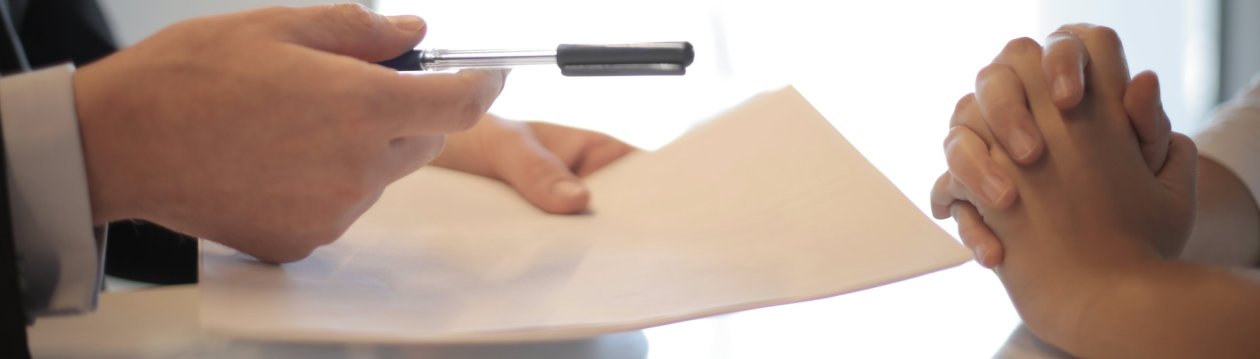 One person holds a pencil over a piece of paper while another person sits across from them with their hands clasped. One person holds a pencil over a piece of paper while another person sits across from them with their hands clasped.