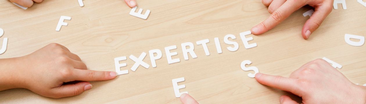 Several hands point to white letters on a wooden table spelling the word “EXPERTISE”.