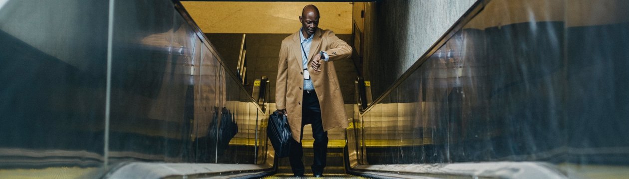 Ein Mann in einem beigefarbenen Mantel und mit einer Aktentasche in der Hand steht auf einer Rolltreppe und schaut auf seine Uhr.