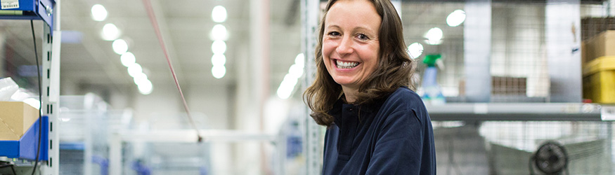 Brunette woman wearing a dark top stands in a warehouse and smiles at the camera.