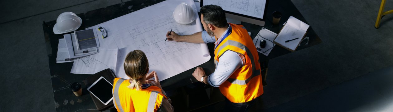 Two people in high-visibility waistcoats bend over a table with construction plans