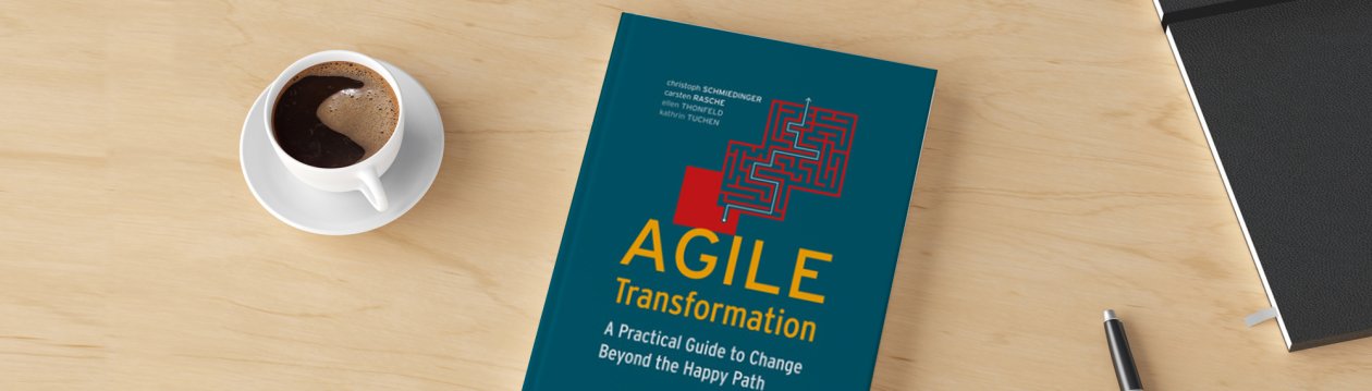 On a table is the book "Agile Transformation", next to it is a cup of coffee, a pen and a notebook.