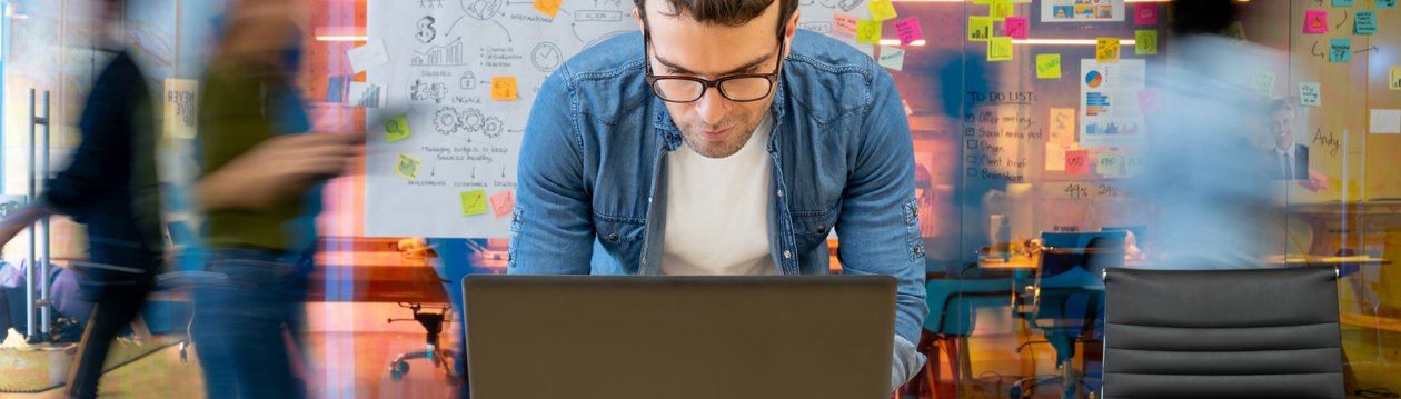 Ein Mann beugt sich über seinen Laptop, im Hintergrund laufen Menschen an einem Whiteboard mit Notizen vorbei. Ein Mann beugt sich über seinen Laptop, im Hintergrund laufen Menschen an einem Whiteboard mit Notizen vorbei.