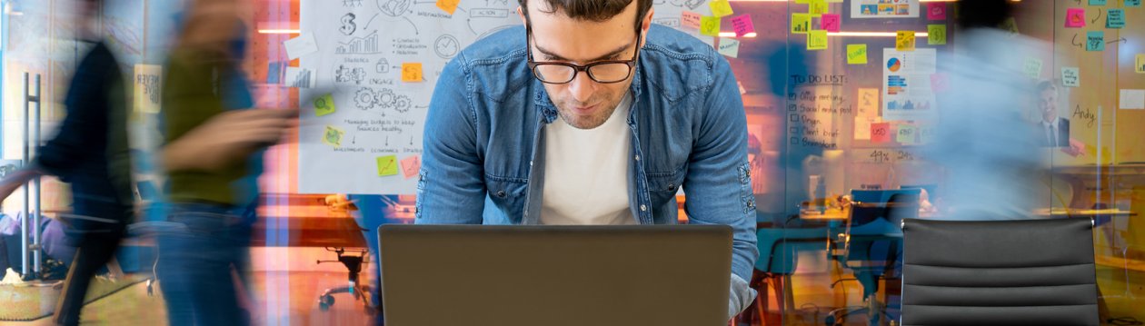 A man bends over his laptop, in the background people walk past a whiteboard with notes.