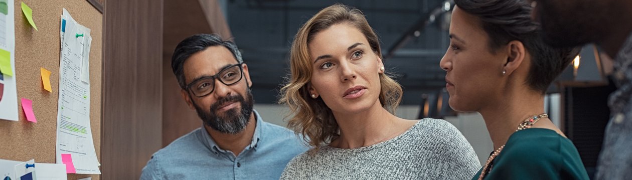 Two women and a man talking in front of a bulletin board