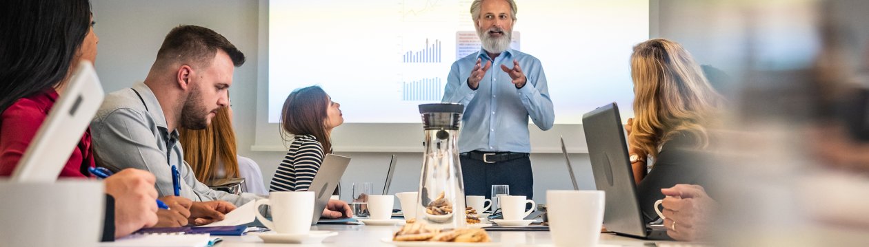 A man presents in front of a screen while several seated people listen at a table. There are cookies and drinks on the table. A man presents in front of a screen while several seated people listen at a table. There are cookies and drinks on the table.