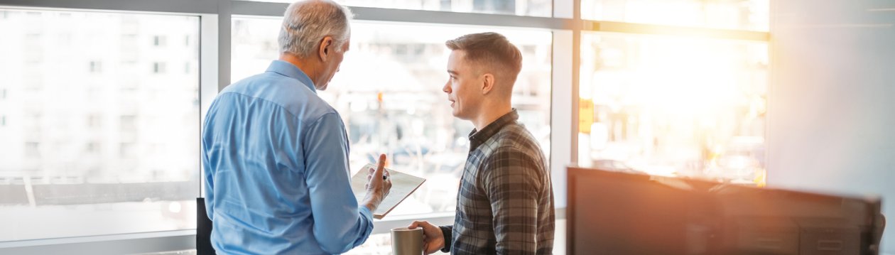 An older man is explaining something to a younger one, which is written on a piece of paper.