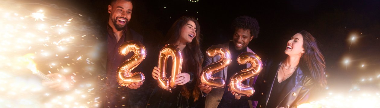 Vier Menschen in einer nächtlichen Silvester-Szenerie, die goldene Luftballons in Form von 2022 halten. Vier Menschen in einer nächtlichen Silvester-Szenerie, die goldene Luftballons in Form von 2022 halten.
