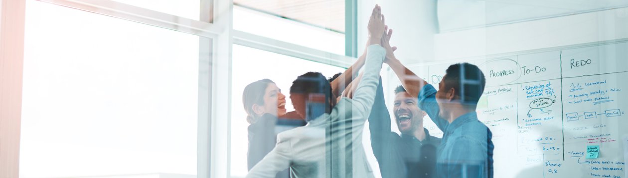 Four business people stand in an office and high-five each other laughing