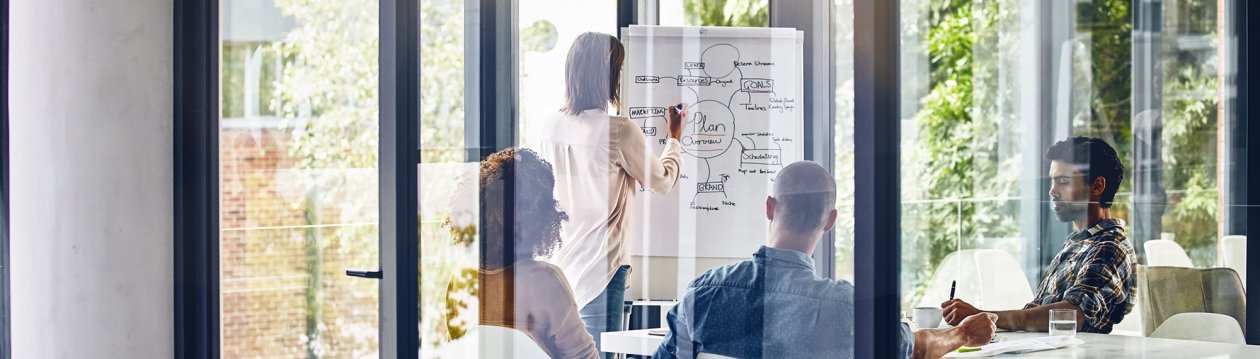 A woman is standing in front of a flipchart showing a marketing plan. 3 other people are watching her write. A woman is standing in front of a flipchart showing a marketing plan. 3 other people are watching her write.