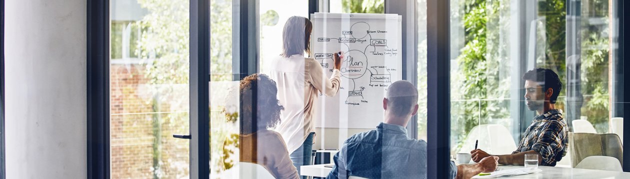 A woman is standing in front of a flipchart showing a marketing plan. 3 other people are watching her write.