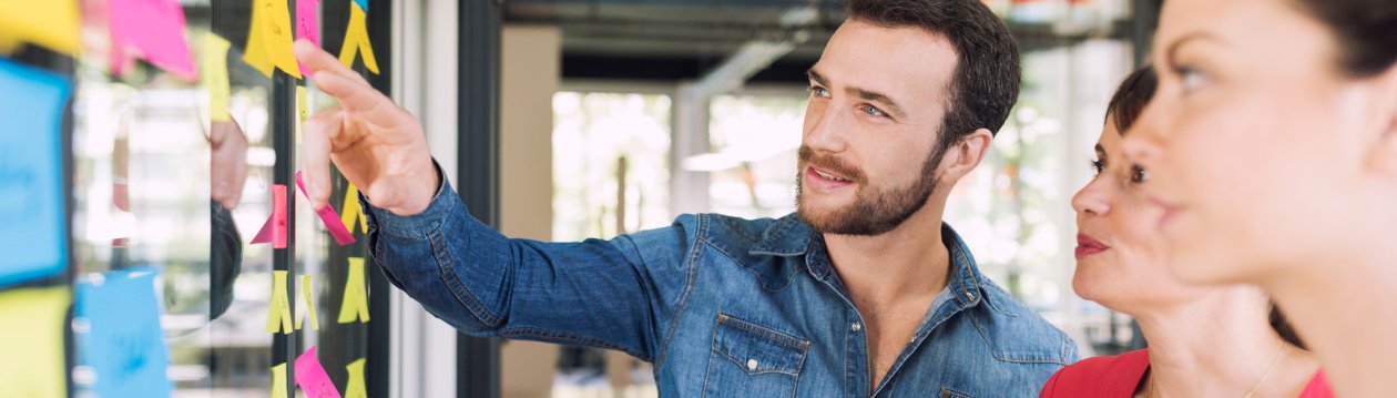 Ein Mann zeigt auf ein Board mit Post-ist, zwei Frauen sehen ihm zu.