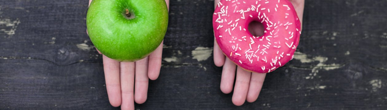 Zwei Hände, von denen eine einen grünen, gesunden Apfel und die andere einen pinken, ungesunden Donut hält. Zwei Hände, von denen eine einen grünen, gesunden Apfel und die andere einen pinken, ungesunden Donut hält.