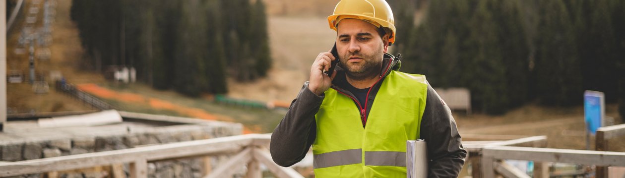 A man in a high-visibility vest and helmet is on the phone at a construction site.