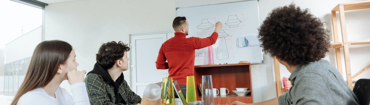 Ein Mann mit rotem Pullover zeichnet UFOs auf ein Whiteboard, 3 weitere Personen sitzen an einem Tisch und schauen ihm zu. Ein Mann mit rotem Pullover zeichnet UFOs auf ein Whiteboard, 3 weitere Personen sitzen an einem Tisch und schauen ihm zu.