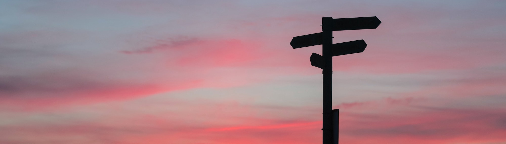 Ein Pfosten mit vier Schildern die in unterschiedliche Richtungen zeigen, vor einem Sonnenuntergang.
