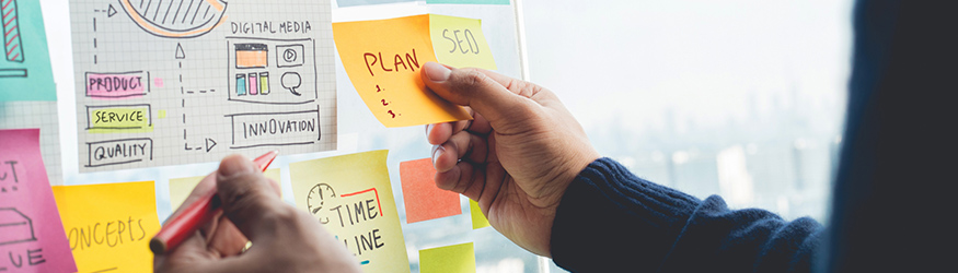 A person attaches a sticky note with the wording "plan" to a pin board.