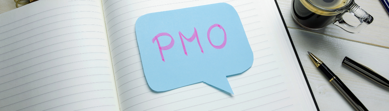 Notizbuch mit Sprechblasen-Zettel „PMO“, daneben Stift und Tasse Kaffee auf einem Tisch.