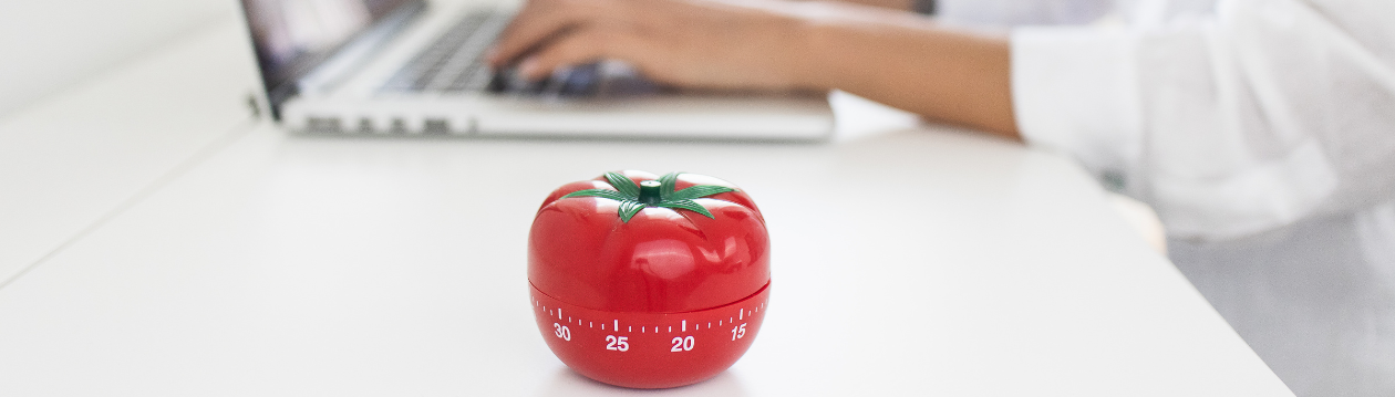 Eine rote Küchenuhr in Tomatenform steht auf einem weißen Tisch vor einer Person, die an einem Laptop tippt. Eine rote Küchenuhr in Tomatenform steht auf einem weißen Tisch vor einer Person, die an einem Laptop tippt.