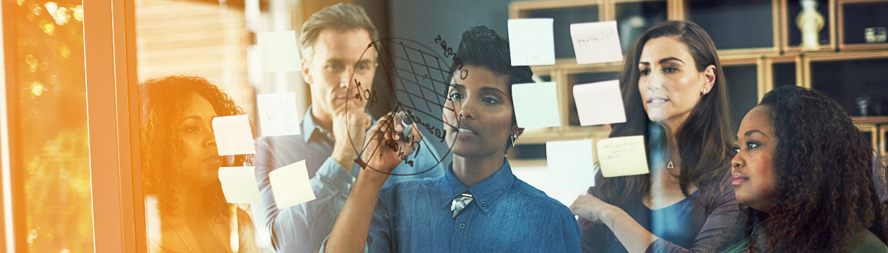 Five people stand in front of a glass wall with sticky notes, one person draws a diagram on it.