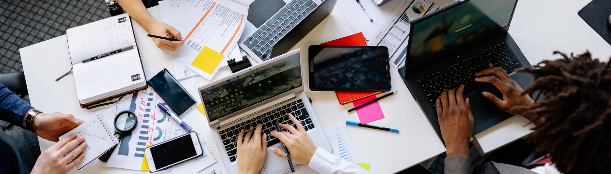 Draufsicht auf Tisch mit Personen, Laptops, Tablet, Smartphones, Notizen, Diagrammen und Haftzetteln während Teamarbeit. Draufsicht auf Tisch mit Personen, Laptops, Tablet, Smartphones, Notizen, Diagrammen und Haftzetteln während Teamarbeit.