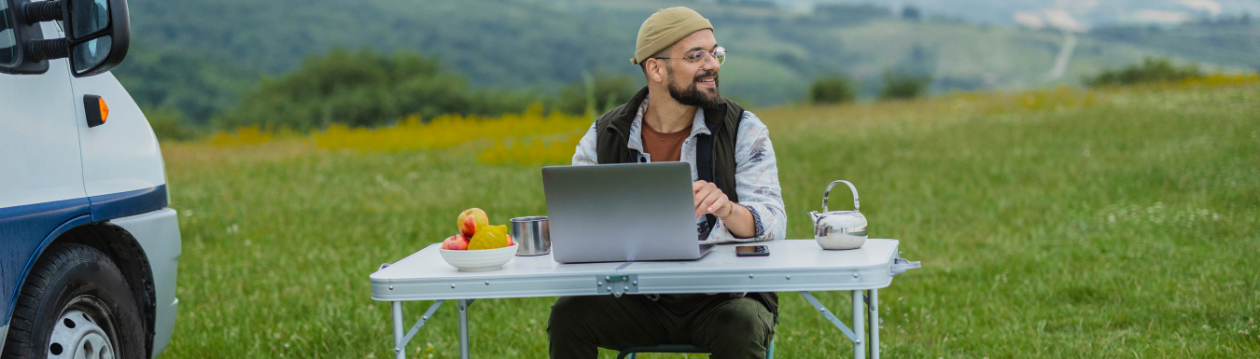Man wearing a beanie sits at a folding table with a laptop beside a van in a grassy field; a bowl of fruit, mug, and teapot are on the table.