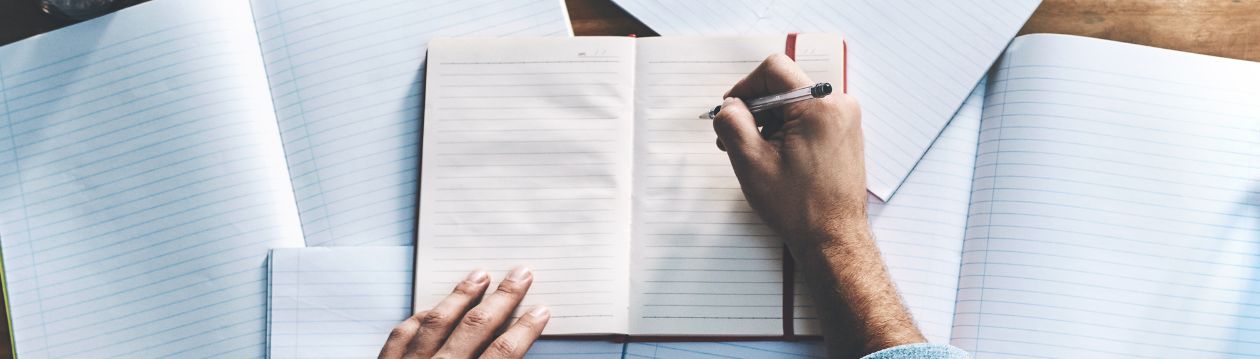 Person schreibt mit einem Stift in ein offenes Notizbuch, umgeben von mehreren linierten Notizheften auf einem Schreibtisch.