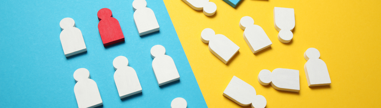 On a surface with a blue and yellow background lie several stylised wooden figures in the shape of people. On the blue side, a single red figure stands amongst white figures and stands out clearly from them. On the yellow side, further white figures lie scattered about, some appearing to have fallen over. The image symbolises themes such as diversity, belonging, exclusion, isolation and group identity. On a surface with a blue and yellow background lie several stylised wooden figures in the shape of people. On the blue side, a single red figure stands amongst white figures and stands out clearly from them. On the yellow side, further white figures lie scattered about, some appearing to have fallen over. The image symbolises themes such as diversity, belonging, exclusion, isolation and group identity.