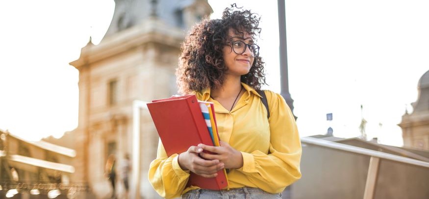 Frau mit lockigem dunklem Haar und Brille, die ein Buch hält und lächelnd in die Ferne blickt. 