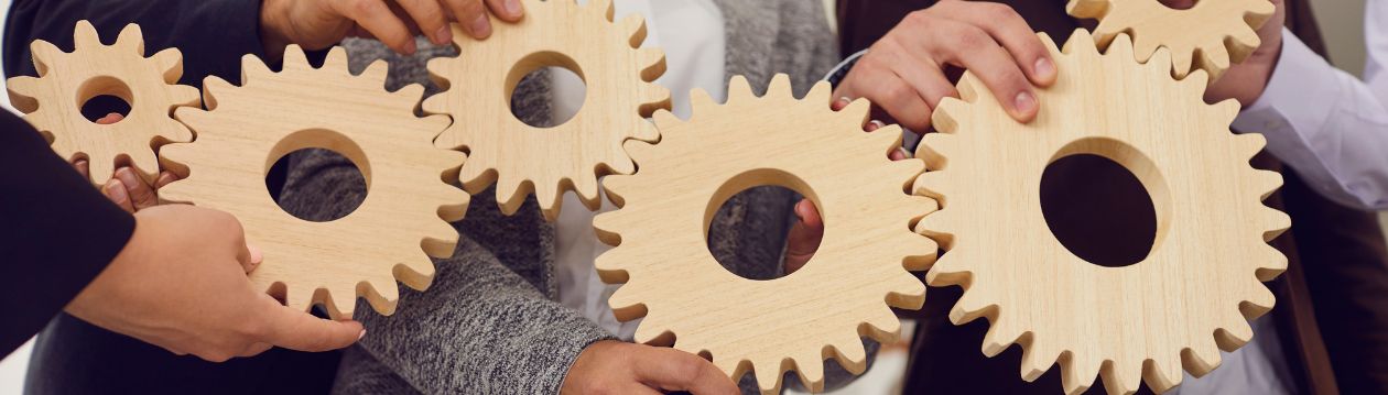 Several people holding and connecting interlocking wooden gears, symbolising teamwork and collaboration.