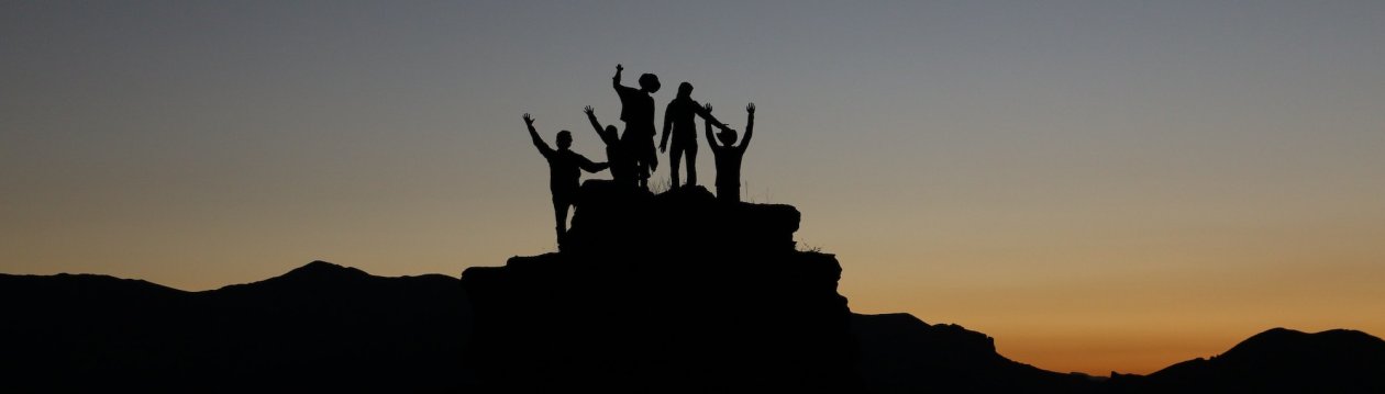 Team Building - Several sillouettes stand on a mountain.