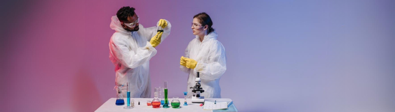 Zwei Wissenschaftler in Arbeitskleidung vor einem Labortisch mit Chemikalien in Reagenzgläsern und einem Mikroskop.